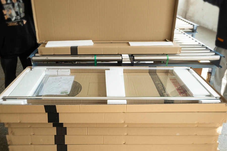 Workers packing tempered glass panels in cardboard boxes with foam padding on a factory conveyor line