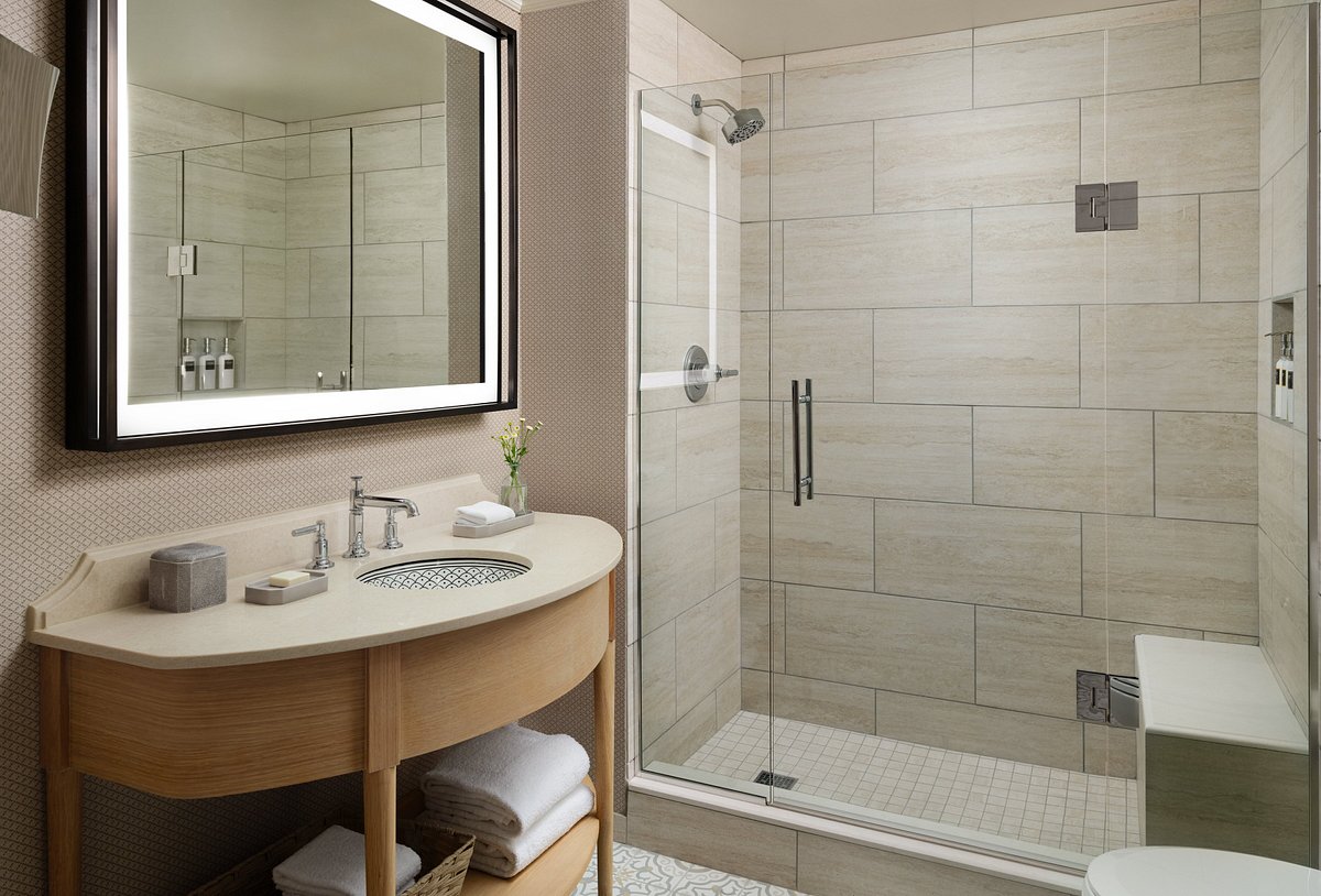 Frameless glass shower door with black handle and built-in marble bench in a beige stone-tiled hotel bathroom