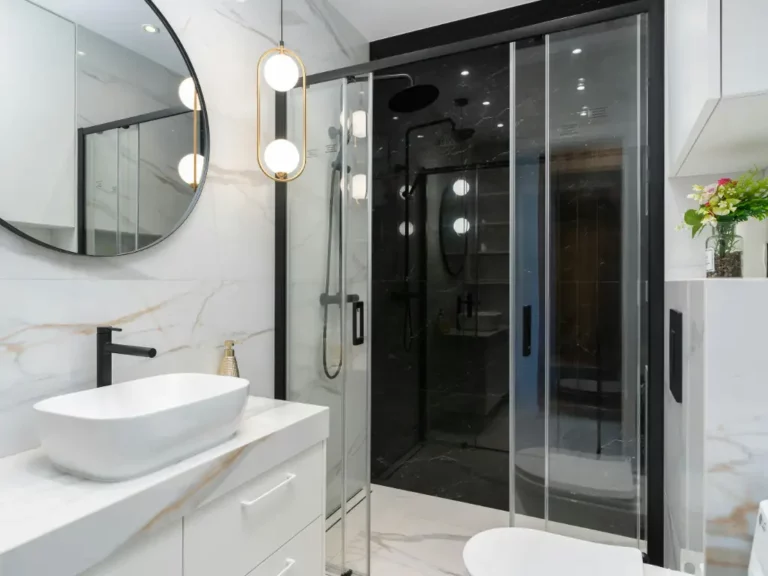 Black framed glass shower door with dark marble interior in a white marble bathroom with round mirror and gold accents