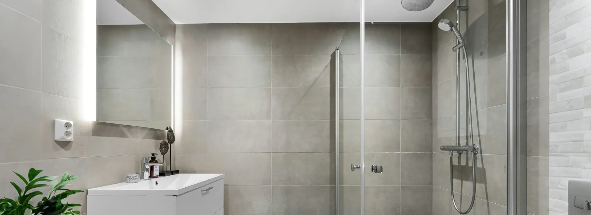 Frameless glass shower door with slim metal frame beside a white vanity and LED mirror in a modern gray-tiled bathroom