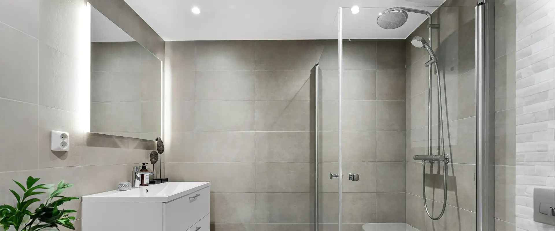 Frameless glass shower screen with chrome handle and rain showerhead in a minimalist gray-tiled bathroom with LED mirror