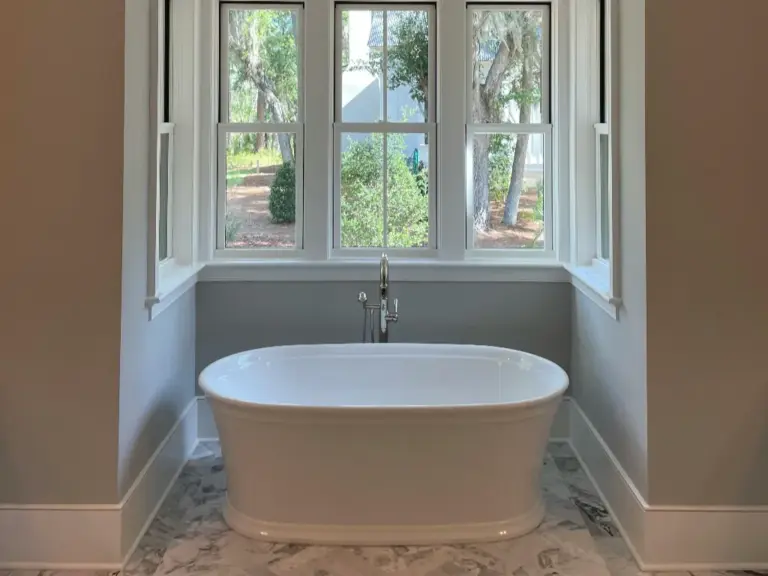 White freestanding bathtub beneath a multi-pane window on a herringbone marble floor in a bright bathroom