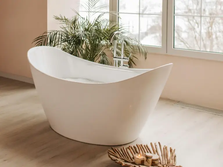 White oval acrylic freestanding bathtub with chrome rim faucet beside a snowy window and green palm plant