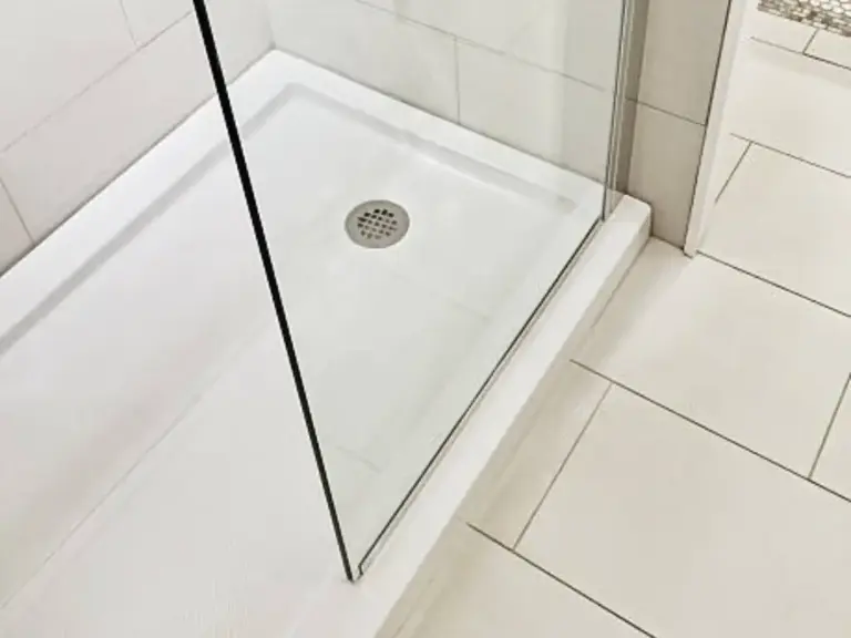 Low-profile white shower base with stainless steel drain and slim black-framed frameless glass panel on light tile floor