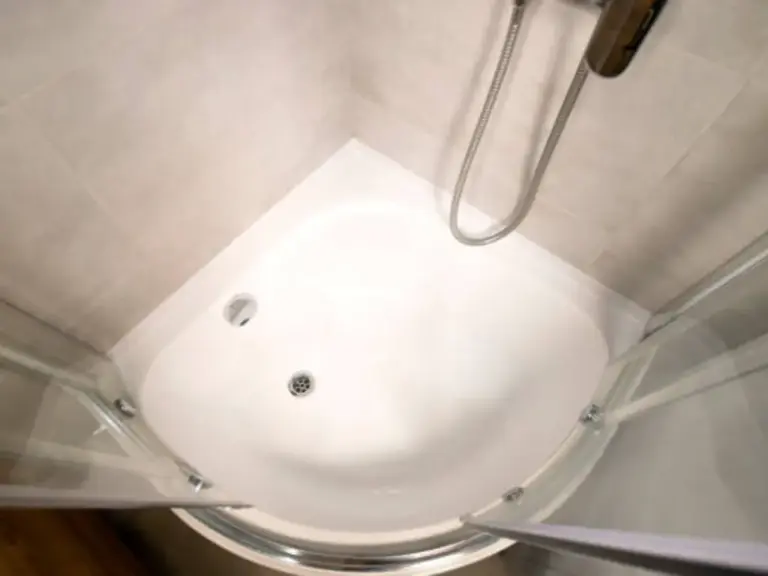 Top-down view of white semi-circular corner shower tray with chrome drain, framed glass doors and beige tile walls
