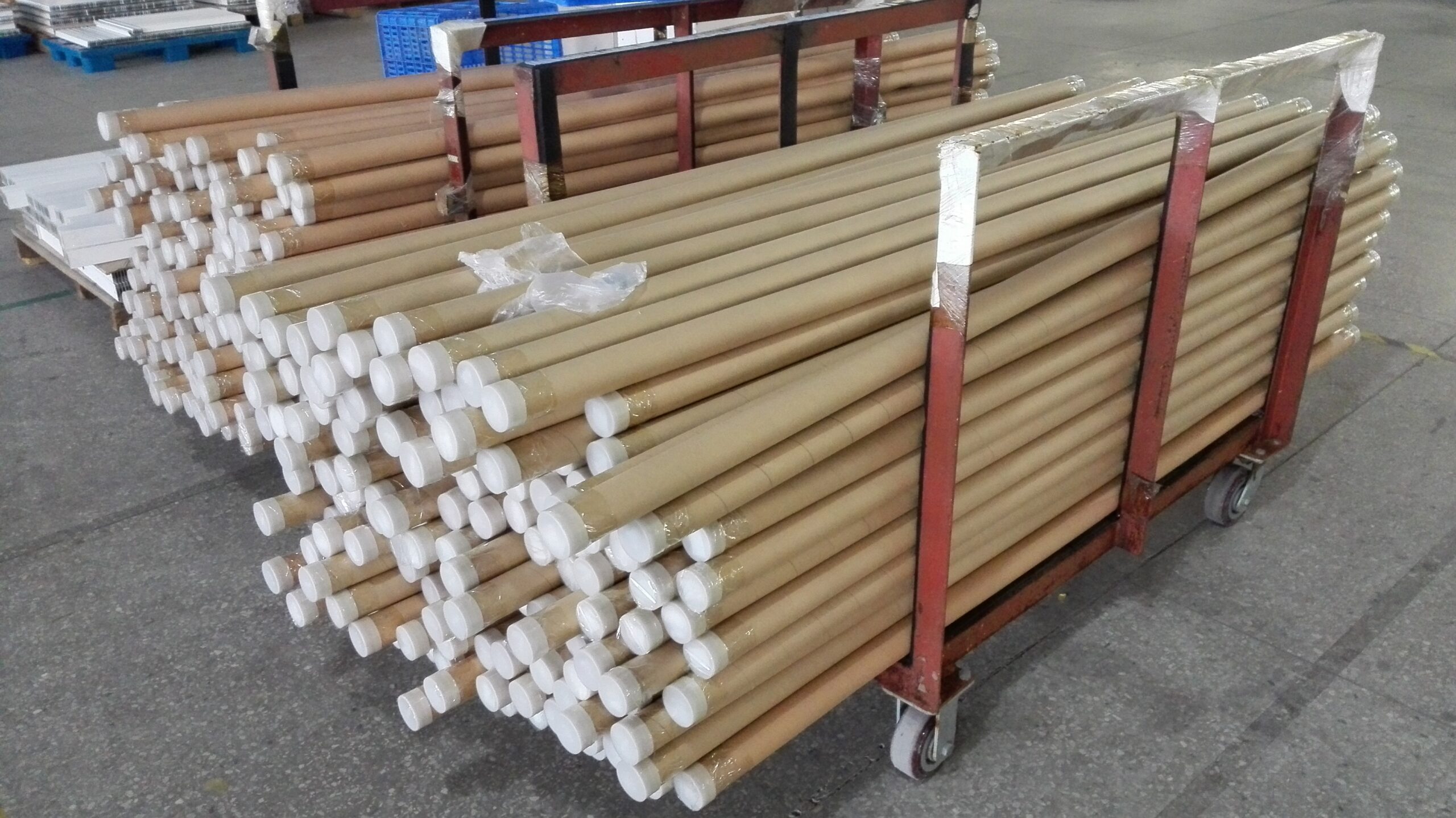 Red metal cart loaded with bundles of brown cardboard packaging tubes staged beside the automatic packaging line